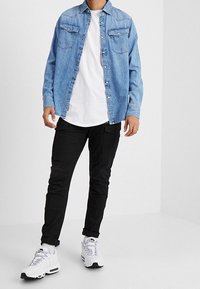 Man som bär en ljusblå denimskjorta över en vit t-shirt, svarta cargobyxor och vita sneakers, stående mot en enfärgad ljus bakgrund.