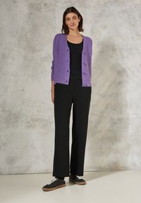 Lila stickad cardigan med knäppning framtill, i kombination med en svart linne och vida svarta byxor, kompletterad av svarta sneakers med vita snören.