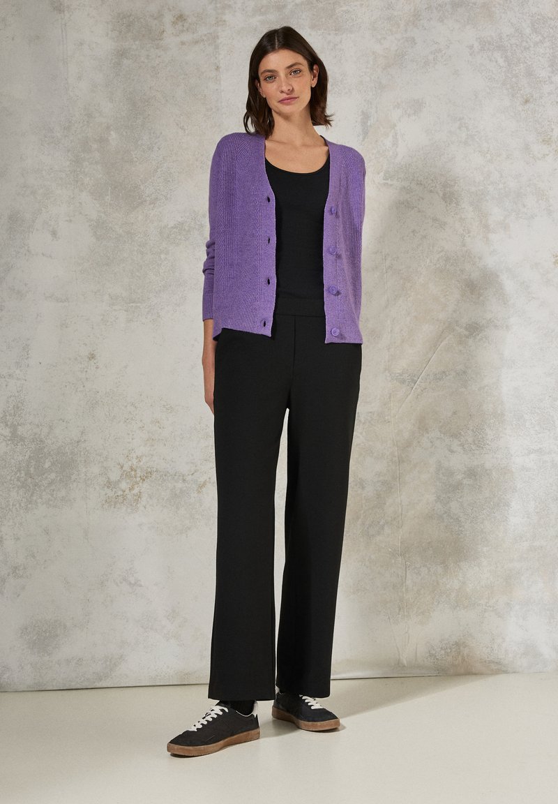 Lila stickad cardigan med knäppning framtill, i kombination med en svart linne och vida svarta byxor, kompletterad av svarta sneakers med vita snören.