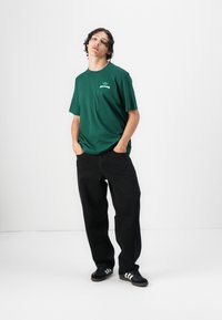 Zöld pamut póló logóval, laza fazonnal, fekete farmernadrággal és fekete sneakers-zel, fehér csíkokkal párosítva. Egyszerű dizájn és hétköznapi stílus.