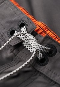 Grijze shorts met een oranje elastische tailleband, voorzien van zwarte gemerkte oogjes en een witte geweven trekkoord. Glad stoftextuur.