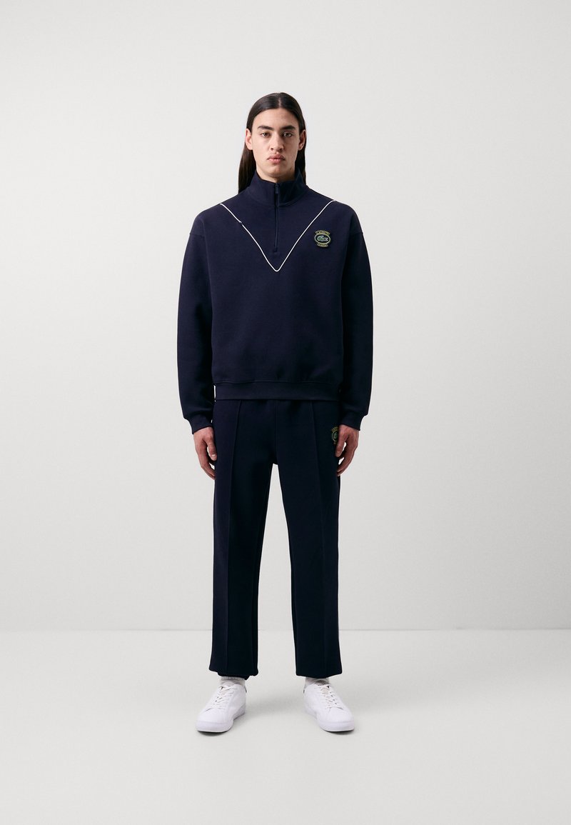 Sweat-shirt marine à col haut et piping blanc, associé à un pantalon assorti. Présente un logo sur la poitrine et des baskets blanches.