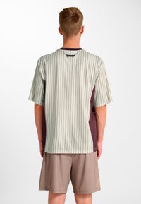 Kurzärmeliges Hemd in Hellgrau mit vertikalen braunen Streifen. Verfügt über weinrote Akzente an den Seiten. Kombiniert mit beigen Shorts.