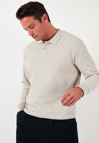 Cămașă polo bej cu mânecă lungă, cu textură moale, manșetă cu trei nasturi și guler contrastant. Pur și simplu cu pantaloni închiși la culoare.