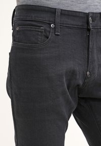 Jeans de mezclilla negra con un corte ajustado, diseño de cinco bolsillos y costuras distintivas. Cierre de botón y cremallera. Textura suave.