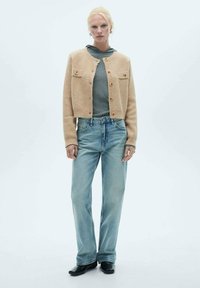 Beige stickad kofta med två bröstfickor, bruna knappar, bärs över en grå hoodie och ljusblå vida jeans. Slät textur, avslappnad passform.