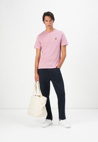Lyserosa bomullst-skjorte med liten gul logo, kombinert med mørk marinebukser og en kremfarget totebag. Jevn tekstur, standard passform.