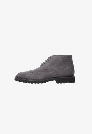 Bottine homme en daim gris avec détails brogue, semelle à crampons noire et fermeture à lacets, inclinée pour montrer le côté extérieur.