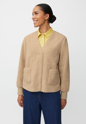 Cardigan beige style peluche avec deux poches frontales, boutons sur le devant, col en V et poignets côtelés, porté sur une chemise jaune et un jean bleu.