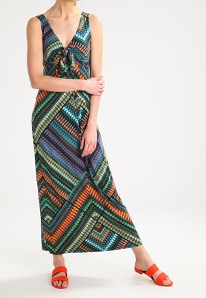 Kobieta w długiej sukience bez rękawów z kolorowym geometrycznym wzorem, z przednim węzłem, nosząca pomarańczowe sandały typu klapki z odkrytymi palcami, stojąca na białym tle.