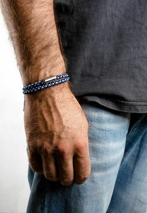 Bratara împletită albastră și neagră cu un închizător metalic, purtată pe un încheietură, cu o mână sprijinită de un picior îmbrăcat în blugi deschiși la culoare.