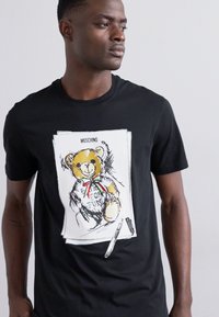 T-shirt en coton noir avec une illustration de teddy bear en cartoon portant un ruban rouge et vert, et le texte "POUR LE PLAISIR" au-dessus de l'ours.