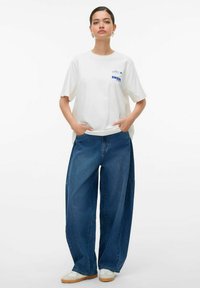 Biały bawełniany t-shirt z niebieskim nadrukiem na piersi, zestawiony z luźnymi niebieskimi spodniami dżinsowymi oraz białymi trampkami z beżowymi podeszwami.