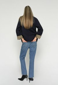 Mujer con cabello largo y liso, lleva una blusa negra con puños de cuadros, jeans azules y botas negras de tacón, de pie con las manos en los bolsillos traseros.