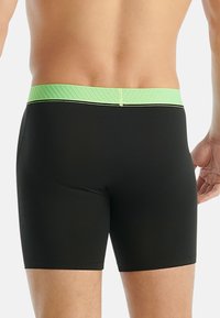 Muži v černých boxerách s zeleným páskem, stojící proti jednolitému světlému pozadí, se zaměřením na dolní část zad a horní části nohou.