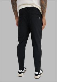 Mustat elastiset vyötäröhousut kapealla leikkauksella, joissa on kaksi takataskua ja pieni logoyksityiskohta oikean takataskun yläpuolella. Yhdistetty mustavalkoisiin tennareihin.