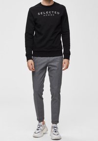 Felpa nera con la scritta "SELECTED HOMME" in grigio, abbinata a pantaloni su misura color grigio chiaro e sneakers bianche con dettagli grigi.