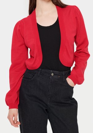 Vrouw draagt een rood kort vest over een zwarte top en zwarte hoge taille jeans, met één hand in de zak, staand tegen een witte achtergrond.