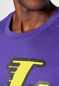 Fialové bavlněné tričko s kulatým výstřihem. Na přední straně má velký černý a žlutý grafický design. Hladká textura a krátké rukávy.