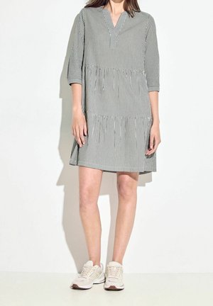 Femme portant une robe rayée verticale noire et blanche à manches trois-quarts et des baskets beiges, debout sur un fond blanc.