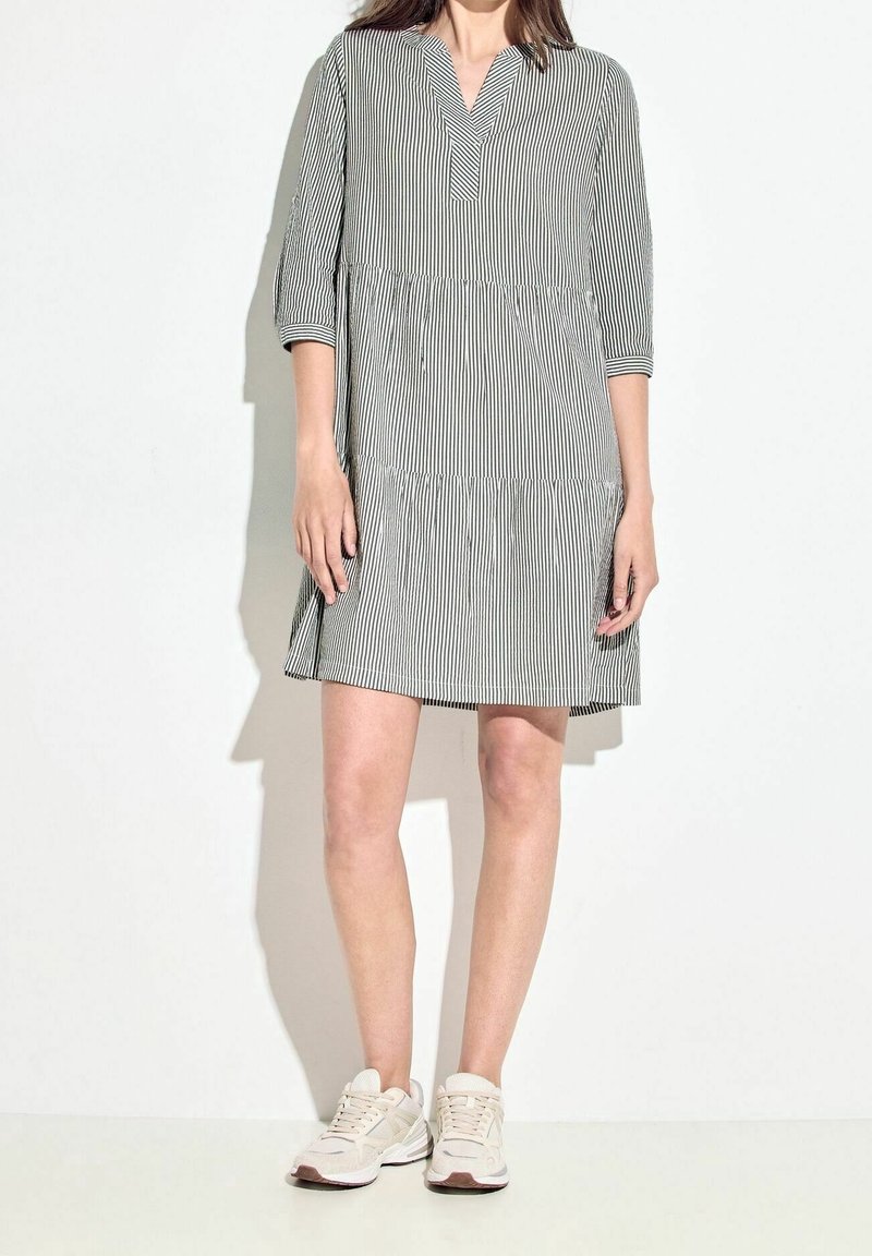 Femme portant une robe rayée verticale noire et blanche à manches trois-quarts et des baskets beiges, debout sur un fond blanc.