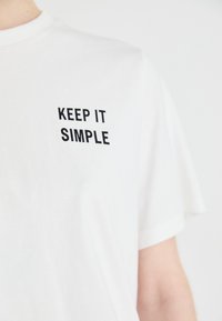 Hvit bomull T-skjorte med korte ermer og rund hals, med "KEEP IT SIMPLE" trykt i fete svarte bokstaver på brystet.