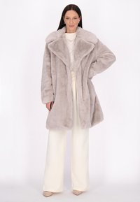 Cappotto in pelliccia sintetica grigio chiaro con ampio colletto, chiusura frontale con bottoni e vestibilità comoda, abbinato a pantaloni crema e tacchi alti. Texture liscia.