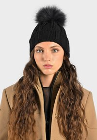 Beanie nera lavorata a maglia con un design a coste spesso e un morbido pom-pom in pelliccia sintetica nera sulla cima. I capelli ondulati di chi la indossa scendono.