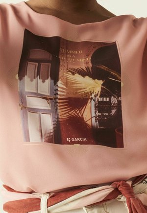 Gros plan d'une chemise rose avec un graphisme de portes, d'ombres et le texte « L'ÉTÉ EST UN ÉTAT D'ESPRIT » ainsi que le nom de la marque « GARCIA » en dessous.