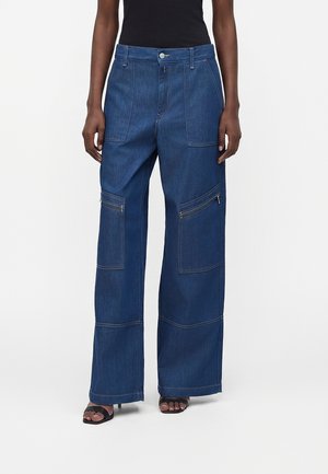 Osoba u širokim tamnoplavim trapericama s više velikih zakrpnih džepova i patentnih zatvarača, uparena s crnim sandalama s trakama.