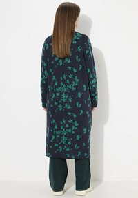 Lång marinblå klänning med gröna blommönster, rundad halsringning och långa ärmar, kombinerad med lösa gröna byxor och vita skor.