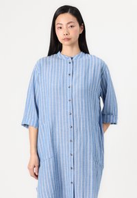 Vestido azul claro de botões com riscas verticais brancas, mangas curtas, gola e dois bolsos laterais. Feito de um tecido texturizado.