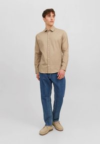 Béžová košile s dlouhým rukávem na knoflíky s límečkem, doplněná modrými džínami a béžovými botami. Košile má hladkou texturu látky.