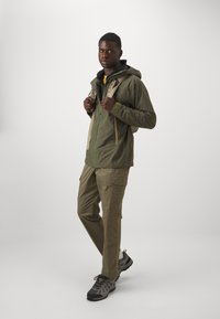 Veste olive avec capuche et poches, associée à un pantalon cargo clair. Chaussures de randonnée grises avec des semelles texturées et des sangles de sac à dos visibles.