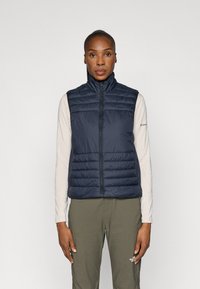 Colete acolchoado azul-marinho com gola alta, apresentando costuras horizontais e fecho de correr à frente. Usado sobre uma blusa de manga longa leve e calças verdes-oliva.