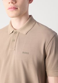 Beige színű póló, textúrált anyagból, két gombos gallérral és hímzett "BOSS" logóval a bal mellen. Hétköznapi stílus.