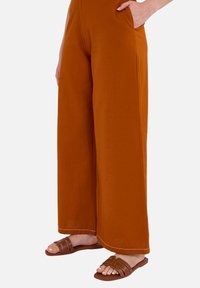 Wijde broek in een roestkleur, gemaakt van een gladde stof. Voorzien van zijzakken en een contrasterende witte stiksels aan de zoom. Bruine slip-sandalen.