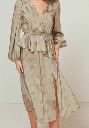 Femme portant une robe portefeuille beige à motifs, avec de longues manches bouffantes, une ceinture froncée à la taille et une fente avant dévoilant sa jambe.