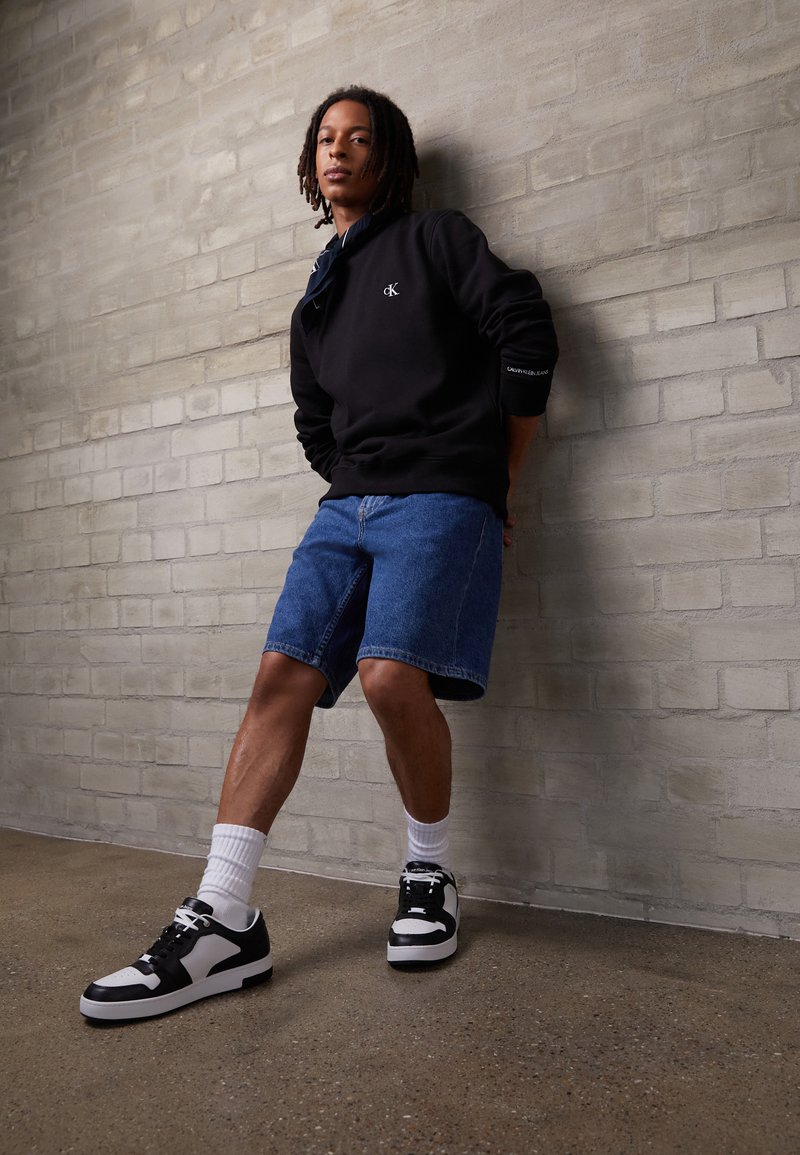 Giovane con dreadlocks che indossa una felpa nera Calvin Klein, pantaloncini di jeans blu, calzini bianchi e sneakers bianche e nere, in piedi contro un muro di mattoni grigio.