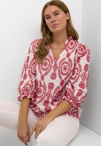 Weiße Bluse mit roten, gestickten Paisleymustern; hat einen V-Ausschnitt, Puffärmel mit gerafften Manschetten und ein leichtes Gewebe.
