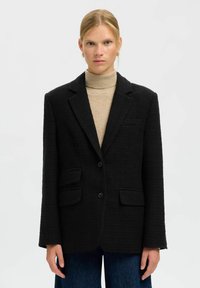 Melnā žakete ar fakturētu audumu, vienrindas aizdare, divas priekšējās kabatas un ar šķautņotu apmali. Kopā ar bēša krāsas augstu apkakli.