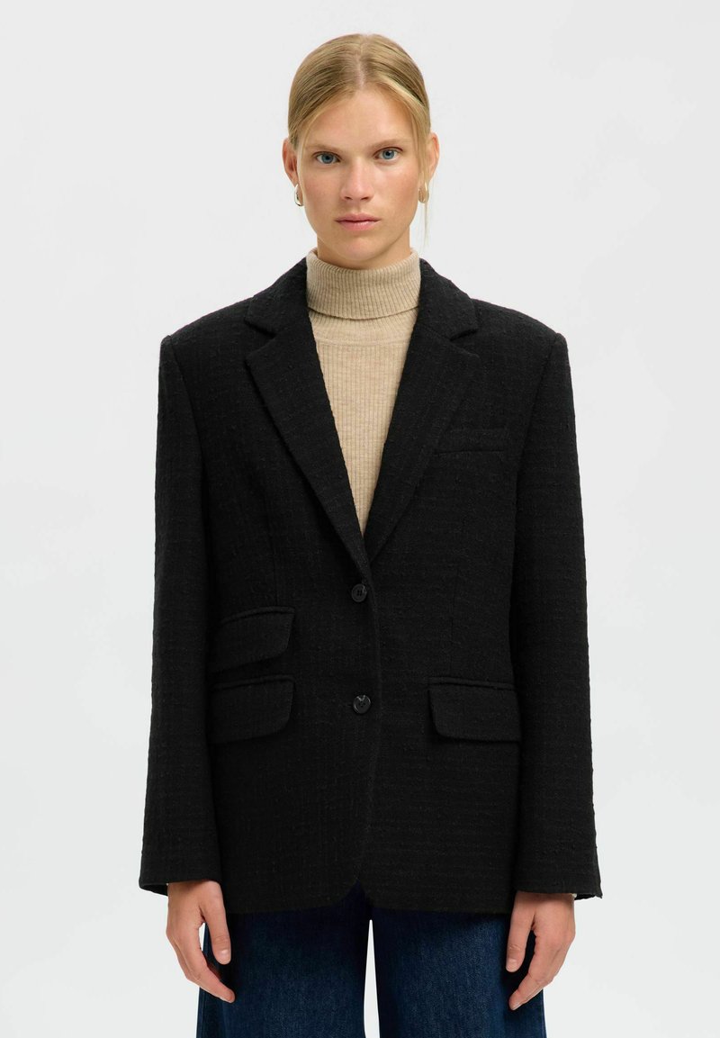 Melnā žakete ar fakturētu audumu, vienrindas aizdare, divas priekšējās kabatas un ar šķautņotu apmali. Kopā ar bēša krāsas augstu apkakli.