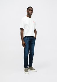 Λευκό βαμβακερό t-shirt με κόκκινο κείμενο, σε συνδυασμό με σκούρες μπλε τζιν και πράσινα αθλητικά παπούτσια ψηλά. Το μοντέλο στέκεται σε ουδέτερο φόντο.