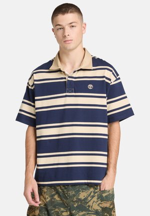 Ung mann iført en rugbyskjorte med marineblå og beige striper og grønne kamuflasjebukser med store lommer, stående med den ene hånden i lommen.