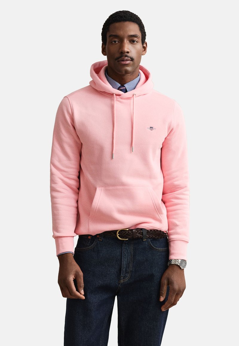 Homme portant un sweat à capuche rose par-dessus une chemise à carreaux bleue avec une cravate sombre, un jean foncé, une ceinture tressée et une montre-bracelet, debout devant un fond blanc.