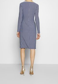 Abito a maniche lunghe in navy con un motivo geometrico ripetitivo. Il design aderente arriva fino al ginocchio ed è abbinato a sandali dorati con cinturini.