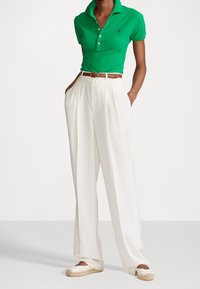 Grön polo med krage, vita veckade byxor, brun bälte och ljusa espadrillos. Tyget är slätt och har en figurnära design.