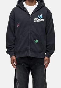 Felpa nera con zip e tasca frontale, decorata con grafiche di farfalle verdi, rosse e blu. Testo "Good for Nothing" sul lato sinistro.
