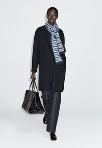 Zwarte wollen jas met een rechte snit, gecombineerd met donkere denim broek. Een blauw-witte gestreepte sjaal voegt textuur toe. Het model draagt een zwarte tote bag.