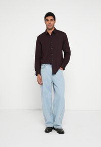 Tmavá vínovočervená košeľa s dlhým rukávom a rovný strih, v kombinácii so svetlomodrými džínsami s voľným strihom a čiernymi topánkami na bielom jednofarebnom pozadí.